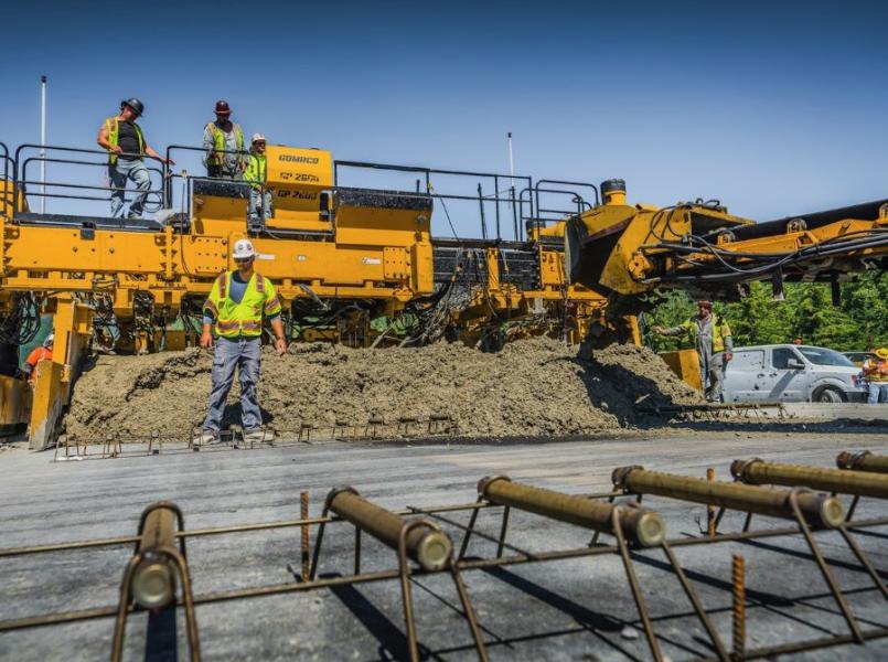 A large yellow paving machine spreads concrete while several workers in safety gear stand around it at a road construction site.