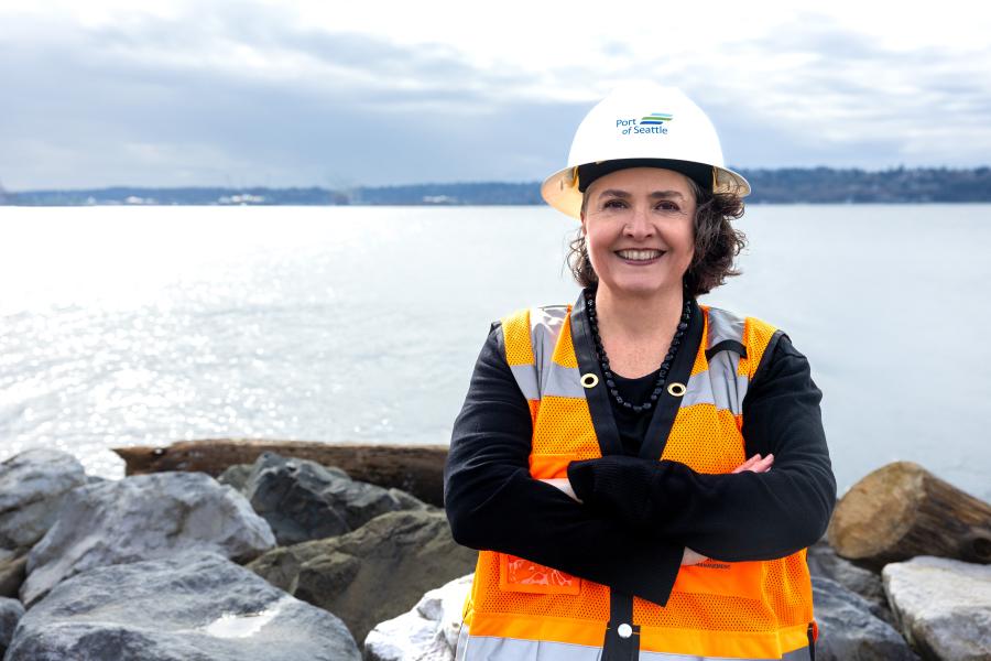 Valerie Huffman wearing a hard hat labeled “Port of Seattle” and an orange safety vest stands with arms crossed on a rocky shoreline, with water and distant land visible in the background.