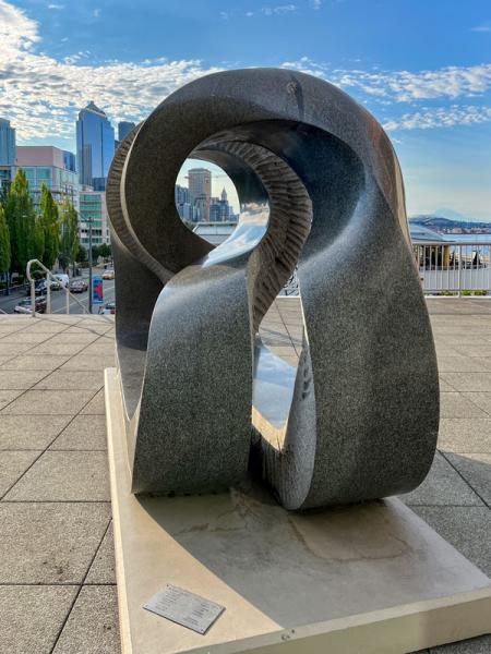 A large abstract stone sculpture with curved openings sits on a plaza overlooking a city skyline and waterfront.