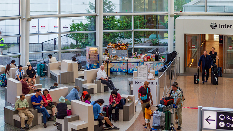 People sit and wait in the Gina Marie Lindsey Arrivals Hall at SEA Airport near International Arrivals, with seating areas, large windows, and a snack stand visible as travelers exit the arrival corridor.
