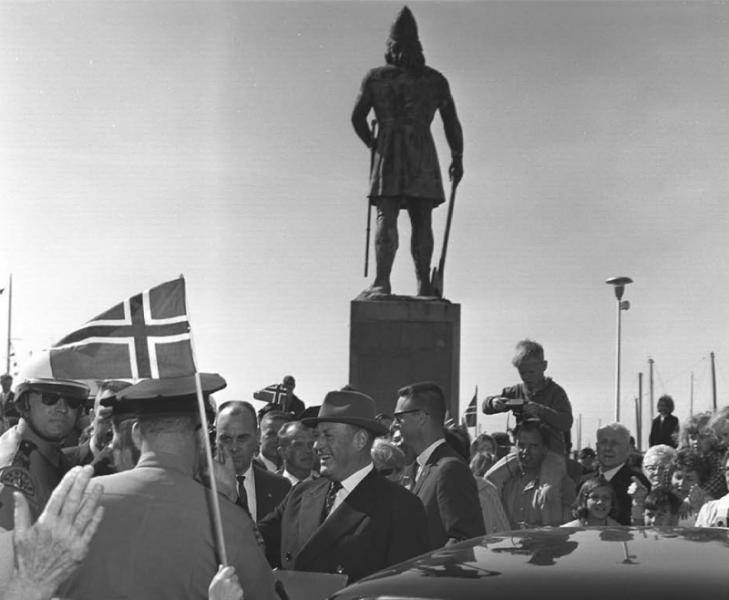 A black‑and‑white photo of a crowd gathered at Shilshole Bay Marina in 1964, with a large statue of a standing figure on a pedestal in the background and people holding flags in the foreground.
