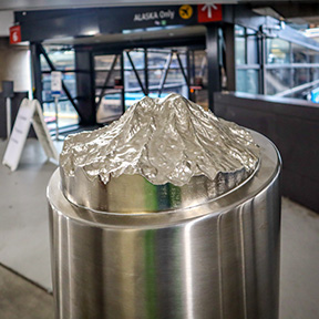 A stainless steel bollard topped with a small, detailed metal sculpture resembling a mountain peak stands inside an indoor walkway area, with glass doors and structural elements visible in the background.
