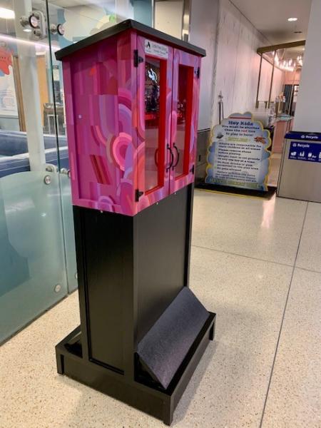 A pink and red swirling‑patterned Little Free Library box stands on a tall black base in an indoor hallway, with a book visible inside through its glass doors.