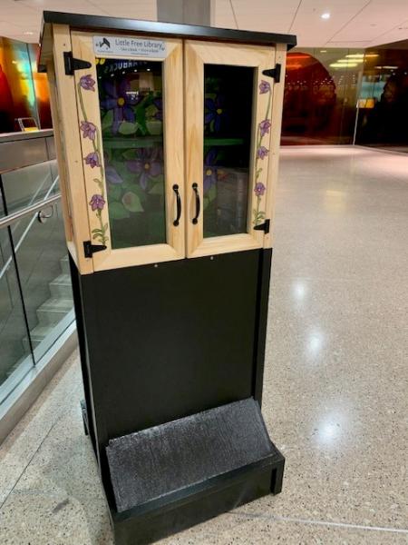 A wooden Little Free Library box with floral designs on the doors stands on a tall black base in an indoor hallway.