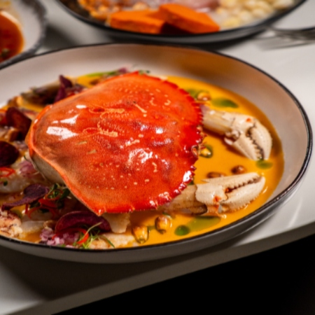 A cooked crab served in a shallow bowl with a rich orange sauce and garnishes.