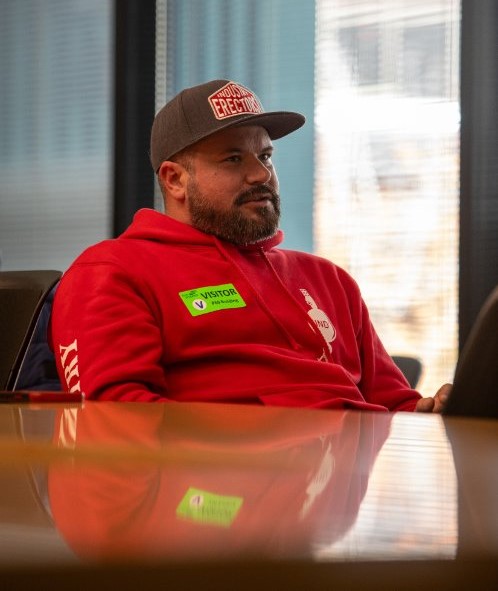 Larry Jacquot in a red hoodie and gray cap with visible text sits at a conference table wearing a green visitor badge.
