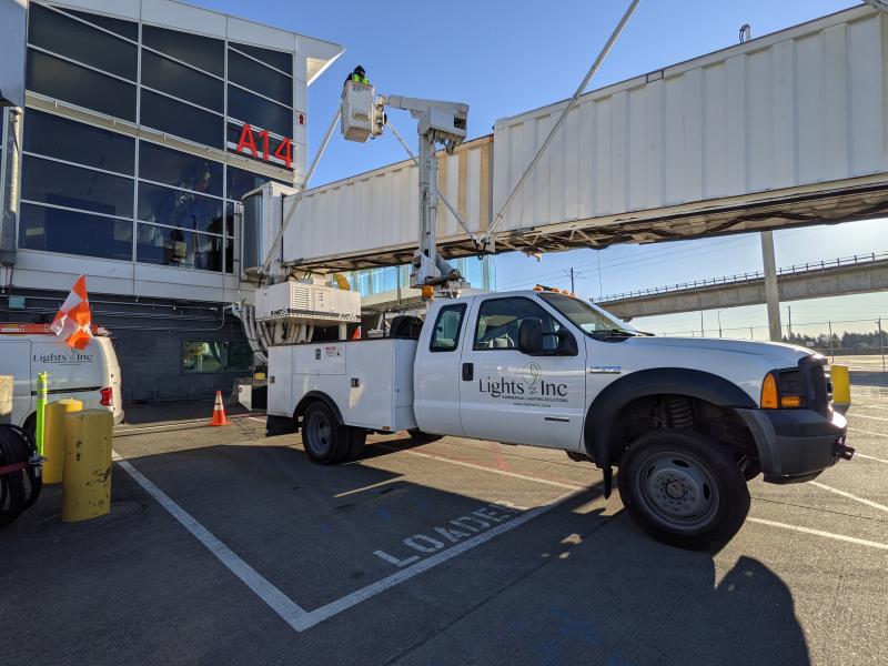 A utility truck with a bucket lift is parked at an airport gate marked A14, where a worker in safety gear is elevated to perform maintenance on the jet bridge.