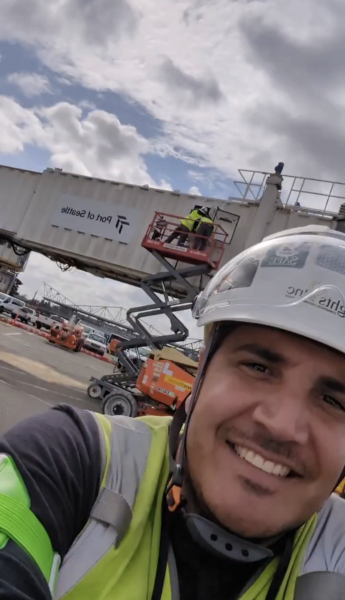 Junior Segura wearing safety gear takes a selfie at an airport worksite, with another worker elevated on a lift near a jet bridge labeled “Port of Seattle.