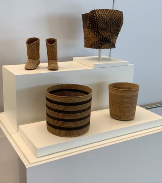 A display of woven objects, including a pair of small woven boots, a patterned woven hat on a stand, and two cylindrical baskets, all arranged on white display platforms.