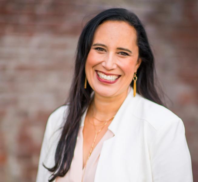 Lisa Keohokaloe Schauer wearing a light-colored jacket and long earrings stands outdoors in front of a blurred brick wall, looking toward the camera.