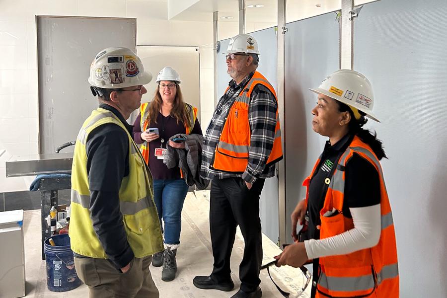Four construction workers wearing hard hats and safety vests stand inside a partially completed building, talking together near unfinished walls and tools.