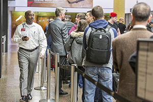 Crowded airport security line with travelers standing behind stanchions while a staff member walks alongside holding a mobile device.