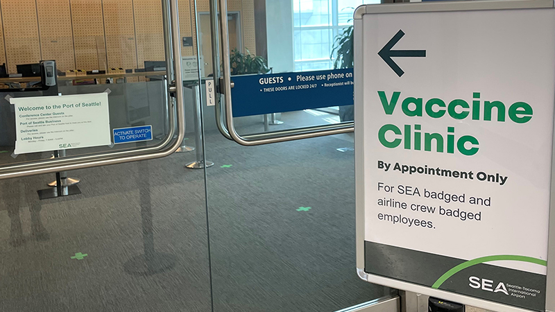 A sign on a glass door directs people to a vaccine clinic for badged employees, with an office area visible inside.