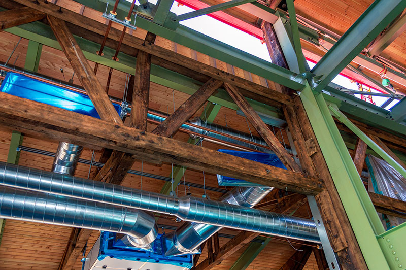 Interior view of exposed timber beams, steel supports, and metal ductwork beneath a wooden roof.