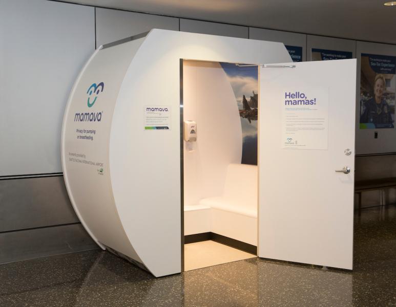 Standalone Mamava nursing pod in an airport hallway, with an open door showing a small private seating area and signage on the exterior.