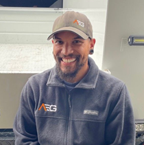 Marcus Cola wearing a cap and a fleece jacket with an AEG logo sits in front of a vehicle and looks toward the camera.