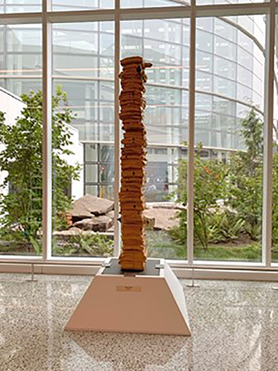 A tall, stacked wood sculpture stands on a white pedestal in a bright atrium with large windows overlooking a landscaped garden.
