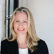 Marilee Hanks wearing a dark blazer and light top is standing indoors near a white wall and window, looking toward the camera.
