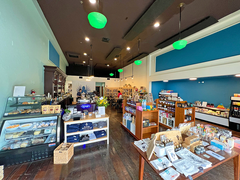 Interior of a specialty shop with display cases of baked goods, shelves of packaged food items, merchandise tables, and a counter at the back, all under hanging green lights.