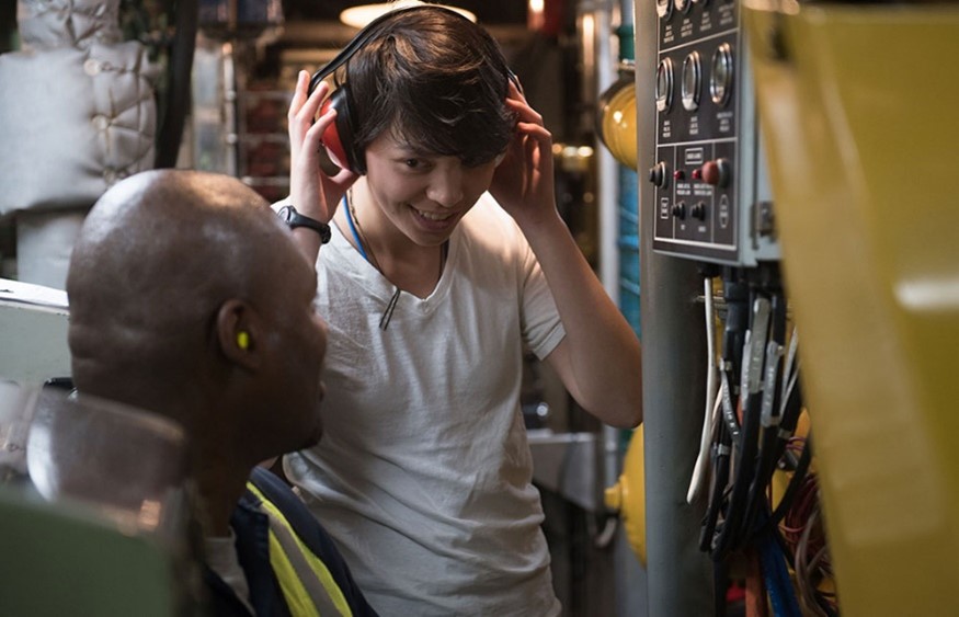 Two individuals in an industrial workspace, with one individual adjusting a pair of over‑ear communication headphones while another individual with earplugs sits nearby.