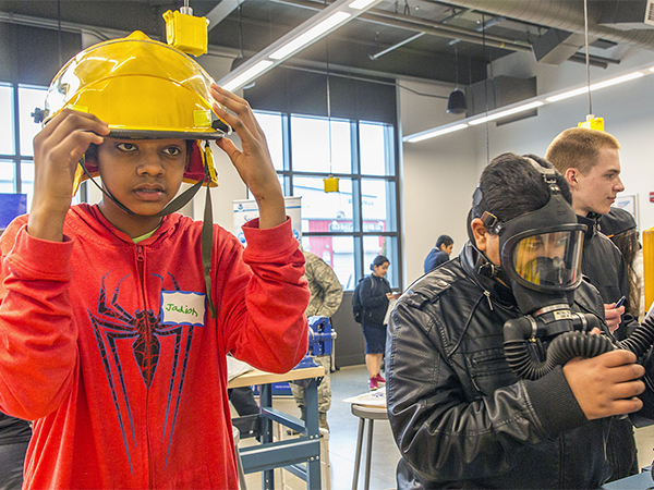 Young man in orange pullover tries on a yellow protective helmet at a Youth Maritime Jobs event.