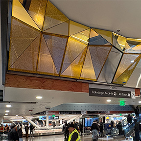 A ceiling installation made of gold and tan triangular panels forms a faceted geometric design above a busy airport concourse.