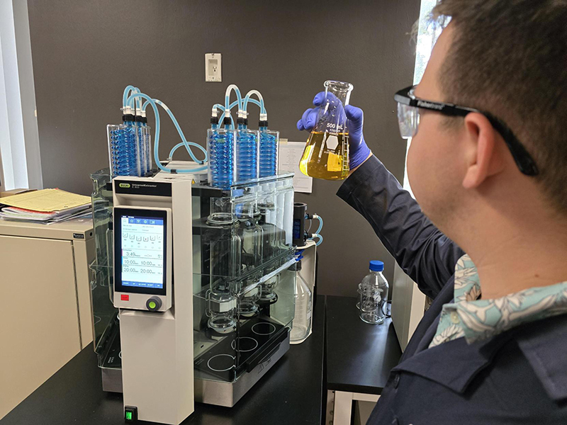 A lab worker holds a flask of yellow liquid near a machine with blue‑filled tubes and a digital display.