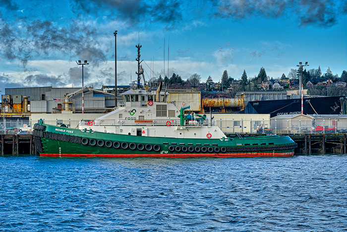 A green and white tugboat named Michael Foss docked beside an industrial waterfront under a partly cloudy sky.