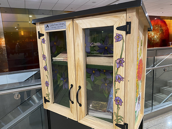 A small wooden Little Free Library cabinet with glass doors and floral artwork on the sides, containing several books and positioned indoors near a railing.