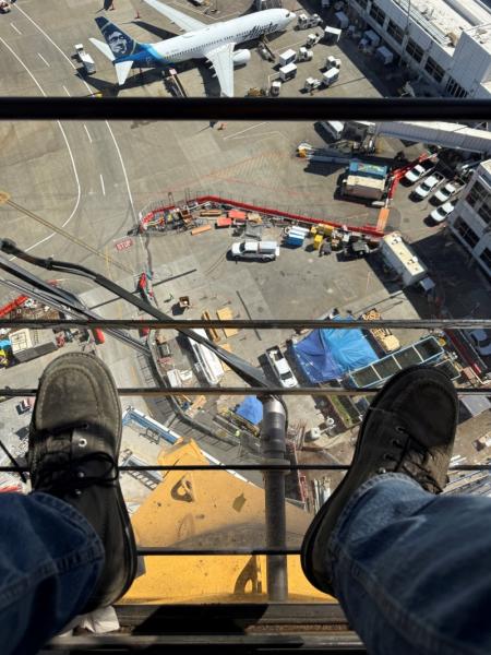 A high view from a tower crane shows the operator’s feet above an airport apron with aircraft, vehicles, and construction activity below.