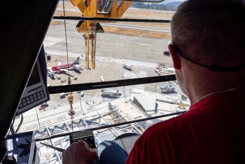 A view from inside a tower crane cab shows an operator looking out over an airport apron with aircraft, vehicles, and construction equipment below.
