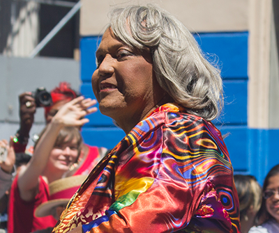 Miss Major wearing a brightly patterned garment participates in an outdoor event, with several people and a blue wall in the background.