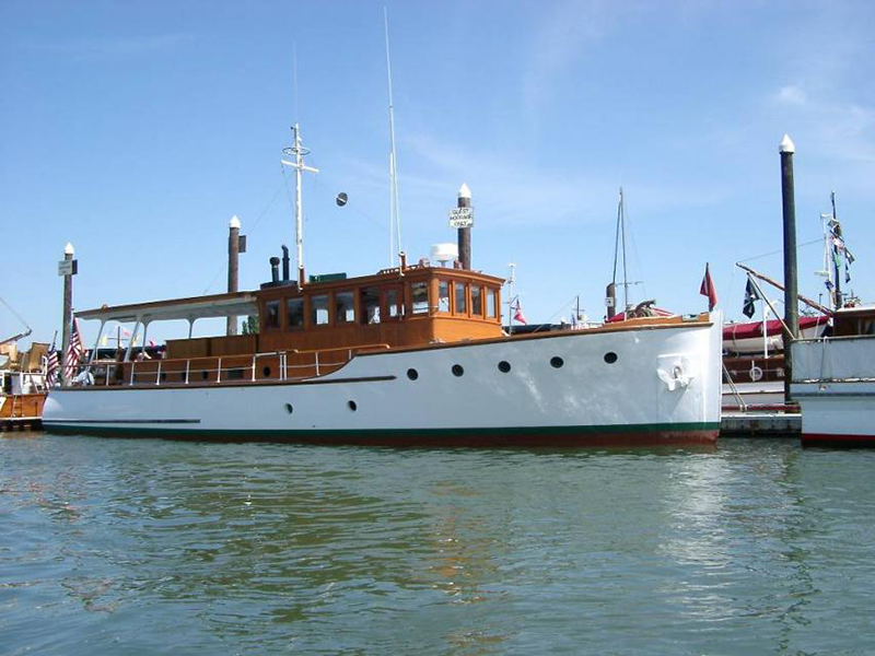 A white boat named “Mitlite” is docked beside other vessels, featuring a wooden cabin structure, open deck area, and several vertical posts along the pier under a clear sky.