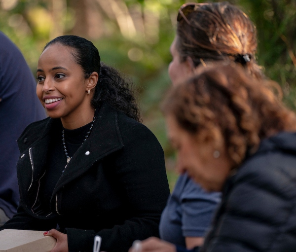A person smiling and talking with others outdoors in a wooded setting.