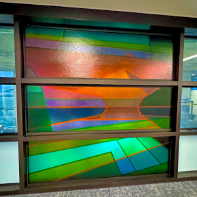 A glass wall artwork by Morgan Madison features layered geometric shapes in vivid colors, including green, blue, red, and orange. The design creates an abstract, angular composition that appears illuminated by light passing through the textured glass panels.