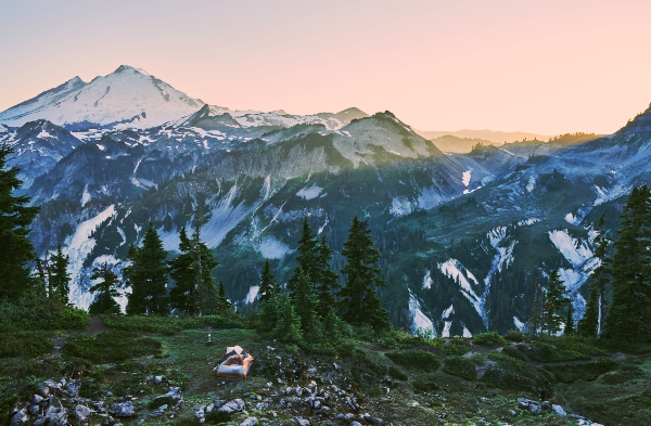 A lone tent sits on a green ridge overlooking rugged, snow‑streaked mountains at sunrise.
