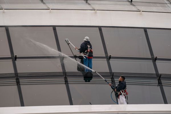 Two workers wearing safety harnesses clean large glass panels on the exterior of a cruise ship using long water-fed poles.