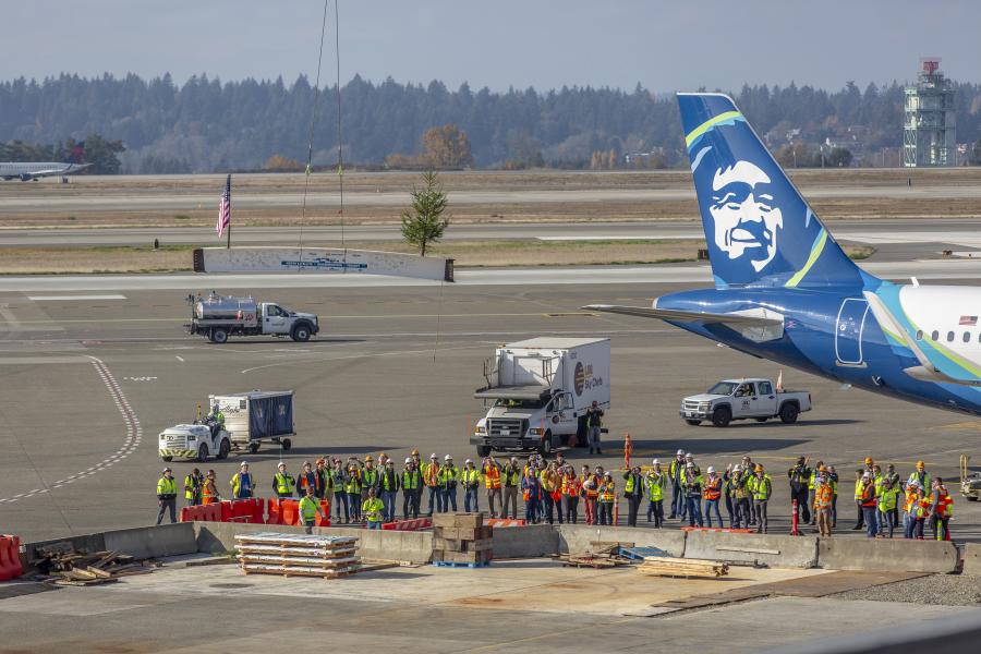 A large group of construction workers in safety gear gather on the tarmac beside an aircraft with a blue tail.