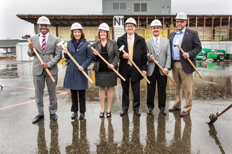 Six people in safety helmets pose with ceremonial sledgehammers at a wet construction site.