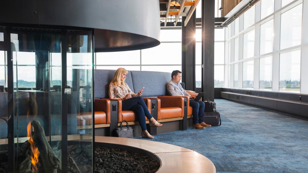 Two people seated in an airport lounge by a glass‑enclosed fireplace, with large windows overlooking the airfield.
