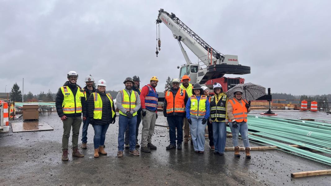 A group of individuals wearing safety vests and hard hats stand together on a construction site near a large crane.