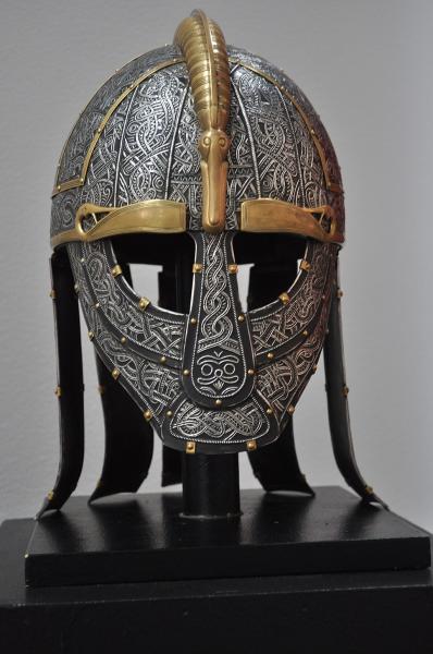 Decorative metal helmet with intricate engravings and patterns, featuring silver-toned etched surfaces, gold-colored accents around the eyes and brow, and a raised central crest. The helmet sits upright on a black display stand against a plain background.