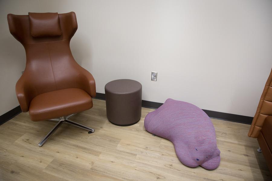 “Comfortable seating area in a Nursing Suite, featuring a modern brown lounge chair, a small round ottoman, and a soft purple floor cushion on a light wood floor.