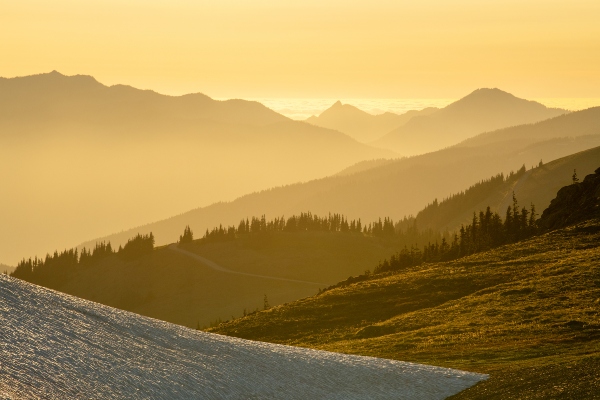 Golden light washes over layered mountain ridges with a patch of snow in the foreground.
