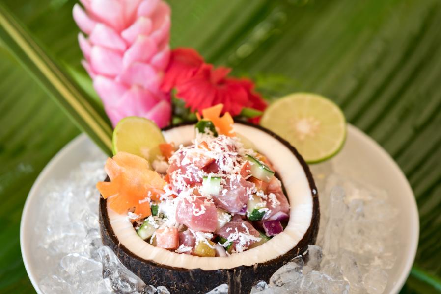 A coconut shell filled with a colorful diced seafood and vegetable dish on ice, with lime slices and flowers in the background.