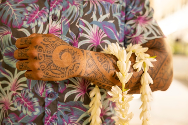 An arm with intricate patterned tattoos holds a strand of white flowers, with a colorful floral shirt visible in the background.