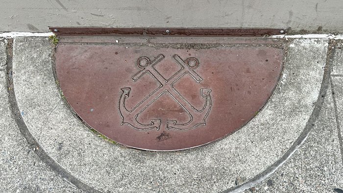 A metal half‑circle plaque embedded in concrete shows an engraved design of two crossed anchors.