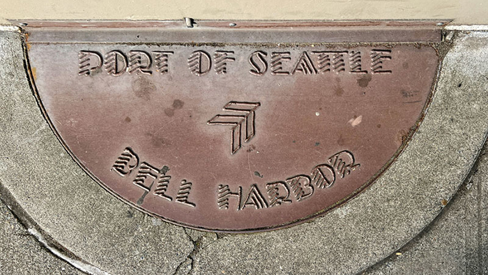 A metal half‑circle plaque embedded in concrete reads “Port of Seattle” at the top and “Bell Harbor” at the bottom, with a stylized arrow‑shaped logo in the center.