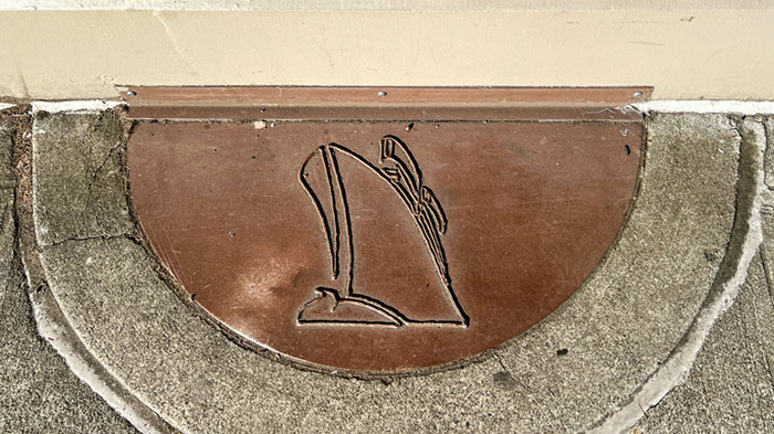 A metal half‑circle plaque embedded in concrete displays a simple engraved outline of a ship’s bow rising from the water.