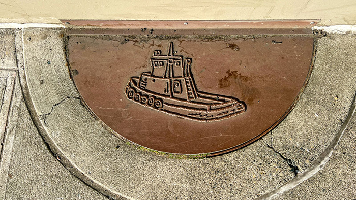 A metal half‑circle plaque embedded in concrete features an engraved outline of a tugboat with a cabin, smokestack, and rounded hull.